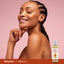 A smiling adult woman relaxes with her eyes closed and chin on hand next to a bottle of Arnica Massage Oil.