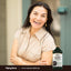 A middle-aged woman laughs with her arms crossed next to a Black Seed Oil bottle.