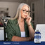 An elderly woman uses a laptop with a Citicoline bottle next to her.