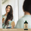Young woman smiles at her reflection while brushing long hair, next to a Castor Oil bottle.
