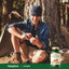 A middle-aged man sits against a tree, looking at his phone, with a Cayenne bottle beside him.