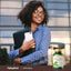 A young adult woman smiles while holding a folder and phone beside a bottle of Gotu Kola.
