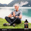 An older man meditates on grass by a lake with a bottle of MCT Oil beside him.