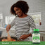 A young adult woman stirs food in a frying pan, with a Whole Greens Fruits & Vegetables bottle nearby.