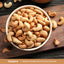 A bowl of organic roasted unsalted cashews on a dark wooden board, with some cashews scattered around.
