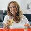 Adult smiling while holding a glass of orange juice, with a Piping Rock Absorbable Vitamin C Plus Zinc, 180 softgels bottle shown in the bottom corner.