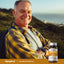 An older man with a backpack smiles on a hillside, holding a bottle of Sea Buckthorn Oil.