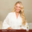 Young woman in white bathrobe smiling with Sweet Almond Oil in hand.