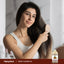 Young woman smiles while brushing her long hair with a wooden brush, Sweet Almond Oil bottle nearby.