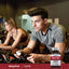 Young adults cycling on stationary bikes with a Taurine bottle nearby.