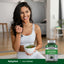 A young adult woman smiles while sitting on the floor and eating a salad, next to a bottle of Acai Supreme.