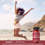 A young woman jumps on the beach with arms raised next to an Energy Booster bottle.