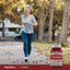 An older woman jogs on a leaf-covered path next to a Vitamin K bottle outdoors.